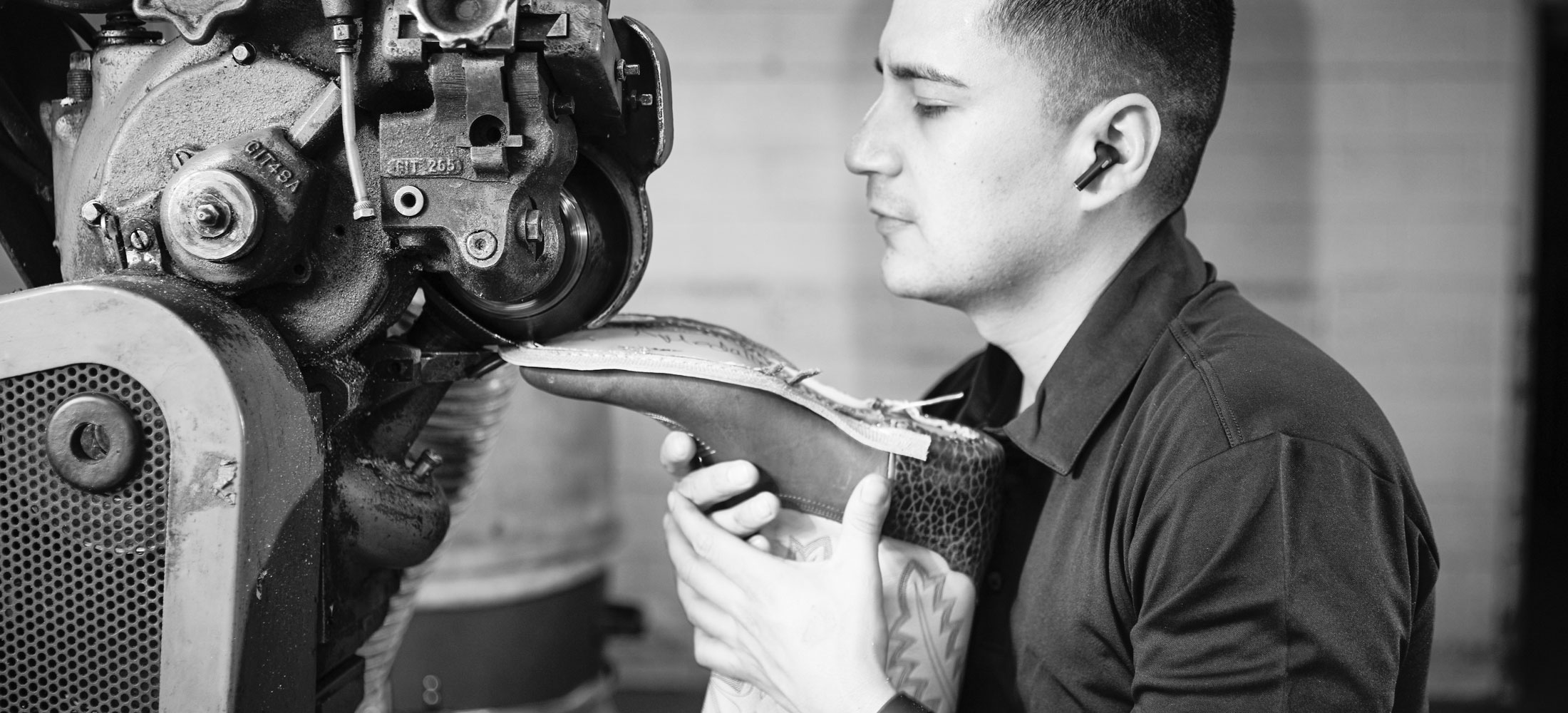 A black-and-white image of a young Hispanic man holding a boot as he’s working on building it.